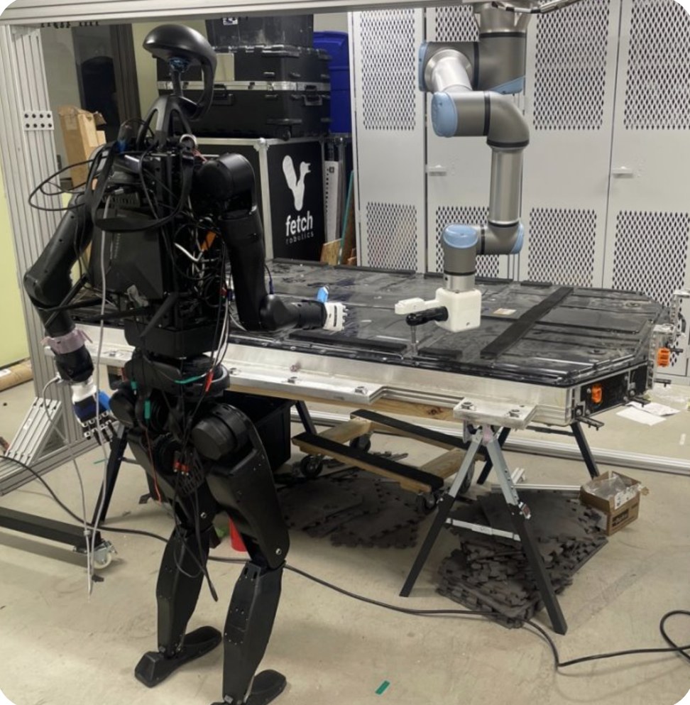 A Unitree H1-2 humanoid standing at a Hyundai Ioniq 5 battery pack beside a gantry-mounted robot arm.